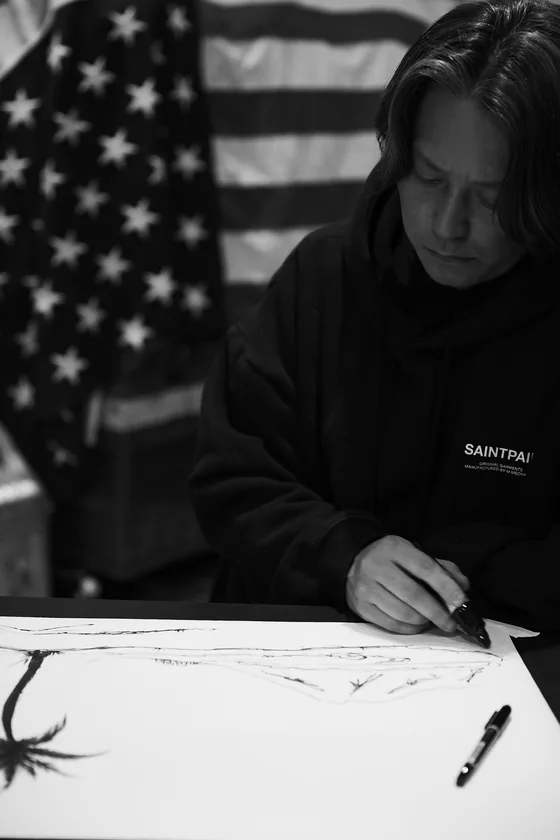 星条旗を背に、紙にペンでヤシの木を描くSHINGIIIIII（モノクロ写真）